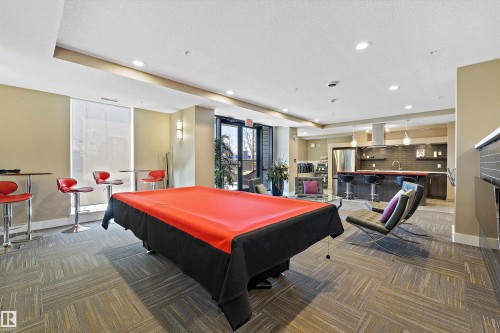 The Recreation Room has a modern kitchen featuring a striking red island countertop, stainless steel appliances, dark wood-finish cabinetry, and a dark subway tile backsplash, with a pool table and a lounge type seating to enjoy with your company. - 307 11080 Ellerslie Rd Sw, Edmonton, AB 