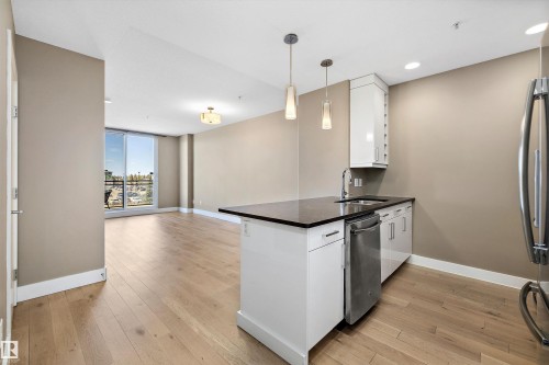 Open-concept living area with wood-finish flooring, a kitchen island featuring a black quartz countertop, and a built-in stainless steel dishwasher. - 307 11080 Ellerslie Rd Sw, Edmonton, AB 