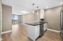 Open-concept living area with wood-finish flooring, a kitchen island featuring a black quartz countertop, and a built-in stainless steel dishwasher. - 307 11080 Ellerslie Rd Sw, Edmonton, AB 