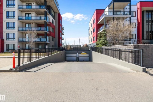 Underground parking access ramp with black metal railings and concrete retaining walls, flanked by two multi-story buildings featuring modern architectural design with red and white facades - 307 11080 Ellerslie Rd Sw, Edmonton, AB 