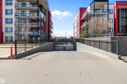 Underground parking access ramp with black metal railings and concrete retaining walls, flanked by two multi-story buildings featuring modern architectural design with red and white facades - 