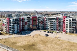 Multi-story residential building featuring a modern facade with red and grey paneling, multiple private balconies, a central entrance, and a paved parking area. - 