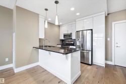Modern kitchen featuring a breakfast bar with dark quartz countertop, stainless steel appliances, white high-gloss cabinetry, recessed lighting, and wood-finish flooring. - 