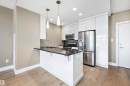 Modern kitchen featuring a breakfast bar with dark quartz countertop, stainless steel appliances, white high-gloss cabinetry, recessed lighting, and wood-finish flooring. - 307 11080 Ellerslie Rd Sw, Edmonton, AB 