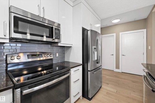 Modern kitchen featuring stainless steel appliances, sleek white cabinetry, a subway tile backsplash, dark countertops, and light wood-finish flooring - 307 11080 Ellerslie Rd Sw, Edmonton, AB 