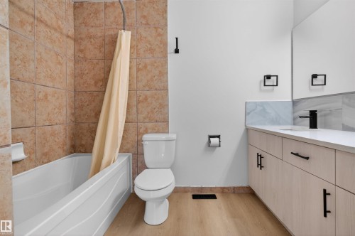 Bathroom featuring a bathtub with tiled surround, a white toilet, a vanity with a light wood-finish base, a white countertop, and black hardware - 7304/7306 78 Avenue, Edmonton, AB 