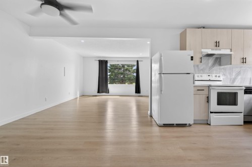 Open-concept living area featuring light wood-finish flooring, white walls, a ceiling fan, and a kitchen with light wood-tone cabinetry, white appliances, and a patterned backsplash - 7304/7306 78 Avenue, Edmonton, AB 