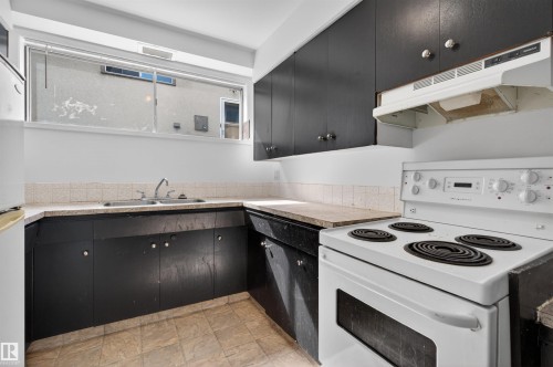 Kitchen featuring a white electric range with an overhead hood, black cabinetry, double basin sink, and a high window - 7304/7306 78 Avenue, Edmonton, AB 