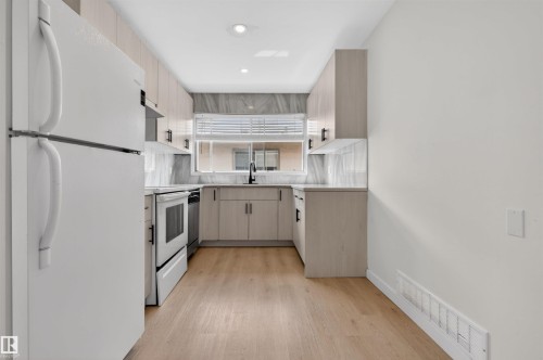 Galley kitchen featuring light wood-finish cabinetry with black pulls, white appliances, and a window above the sink - 7304/7306 78 Avenue, Edmonton, AB 