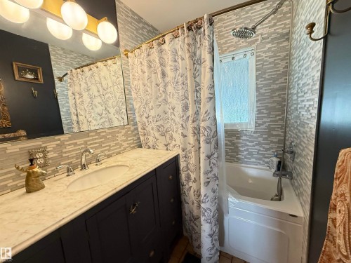 Bathroom featuring a white bathtub, a tiled shower surround, a window with textured glass, a dark vanity with a white countertop and an integrated sink, and a large mirror with overhead lighting - 10912 50 Street, Edmonton, AB 