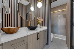 Modern bathroom featuring a vanity with a white countertop and light wood-finish cabinetry, a vessel sink with a gold-finish faucet, and a round gold-framed mirror - 