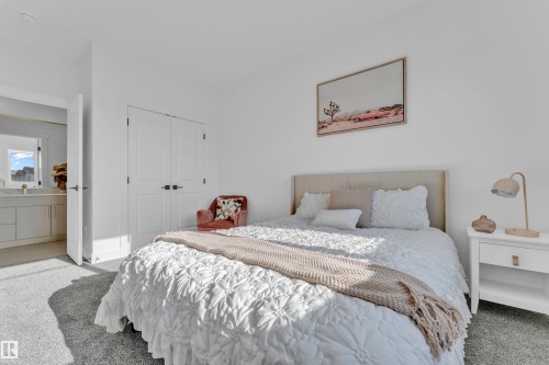 Plush gray carpeted room featuring a neutral upholstered headboard, double-door closet with dark hardware, and a vanity with a mirror and window in the adjacent space - 206 Linden Loop, Leduc, AB 
