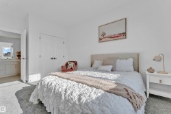 Plush gray carpeted room featuring a neutral upholstered headboard, double-door closet with dark hardware, and a vanity with a mirror and window in the adjacent space - 