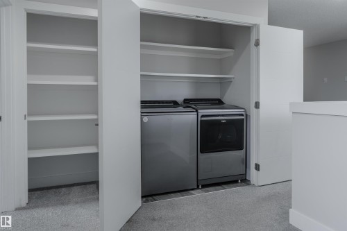 Integrated laundry area featuring a side-by-side washer and dryer, built-in shelving, white bifold doors, and light gray carpeting - 2104 160 Street Sw, Edmonton, AB 