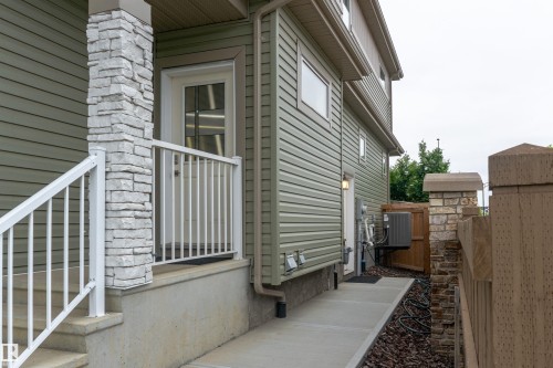 Side exterior featuring light green horizontal siding, stone veneer column accents, a white entrance door with divided lites, and a concrete pathway - 2104 160 Street Sw, Edmonton, AB 