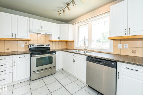 Kitchen featuring white cabinetry, dark countertops, a tile backsplash, stainless steel appliances, and a double basin sink - 7543 149A Avenue, Edmonton, AB 