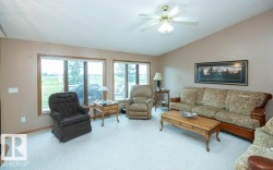 Bright living space featuring large windows with wood-finish trim, a ceiling fan with integrated lighting, and light-colored carpeting - 