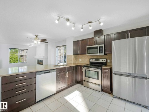 Kitchen featuring dark wood-finish cabinetry, stainless steel appliances, a light-toned countertop, and a tiled backsplash - 77 4050 Savaryn Drive, Edmonton, AB 