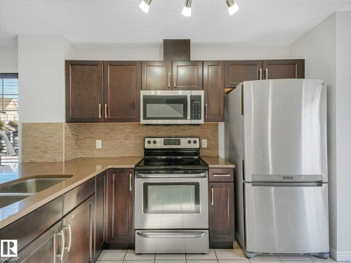 Kitchen featuring dark wood-finish cabinetry, stainless steel appliances, a double basin sink, light-toned countertops, and a tiled backsplash - 77 4050 Savaryn Drive, Edmonton, AB 