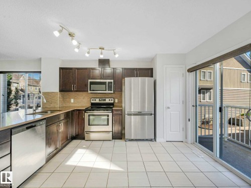 Kitchen featuring stainless steel appliances, dark wood-finish cabinetry, light-toned tile flooring, and a sliding glass door leading to an outdoor patio - 77 4050 Savaryn Drive, Edmonton, AB 