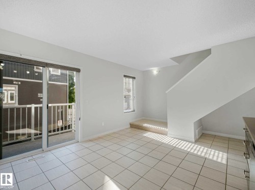 Bright living area featuring white tile flooring, a sliding glass door to a private balcony, a staircase with carpeting, and a window with blinds - 77 4050 Savaryn Drive, Edmonton, AB 