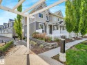 Contemporary townhome exterior featuring gabled roofs, a white picket fence, a dedicated walkway, and mature trees - 77 4050 Savaryn Drive, Edmonton, AB 