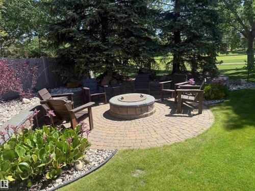 Circular paver patio featuring a built-in fire pit, surrounded by a manicured lawn, established evergreens, and planted garden beds with decorative rock accents - 915 Picard Drive Nw, Edmonton, AB 