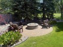 Circular paver patio featuring a built-in fire pit, surrounded by a manicured lawn, established evergreens, and planted garden beds with decorative rock accents - 915 Picard Drive Nw, Edmonton, AB 