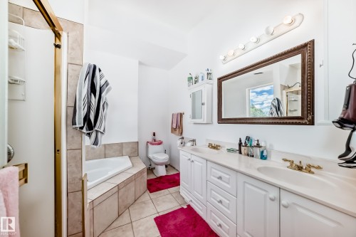 Bathroom featuring a dual vanity with white cabinetry and integrated sinks, a built-in corner bathtub with tiled surround, a separate shower with a gold-toned frame, a wall-mounted medicine cabinet, and a large framed mirror with overhead lighting - 6804 102A Avenue, Edmonton, AB 