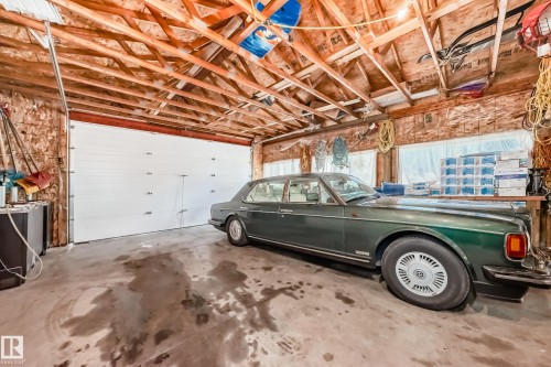 Spacious garage featuring a large white overhead door, exposed wooden ceiling trusses, concrete flooring, windows, and shelving - 6804 102A Avenue, Edmonton, AB 
