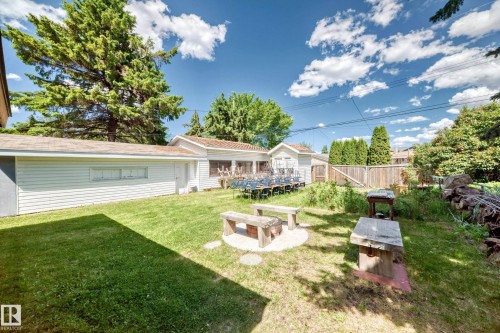 Expansive backyard featuring a manicured lawn, a fire pit area with stone benches, a dedicated outdoor workspace, perimeter fencing, and mature trees - 6804 102A Avenue, Edmonton, AB 