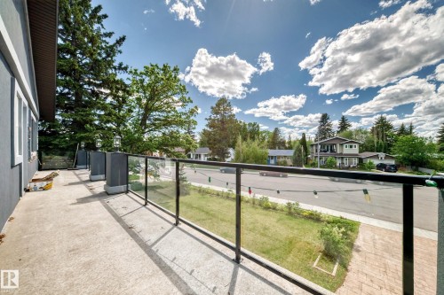 Concrete balcony with a glass and metal railing, featuring a stucco exterior, multiple exterior light fixtures, and a tree-lined streetscape - 6804 102A Avenue, Edmonton, AB 