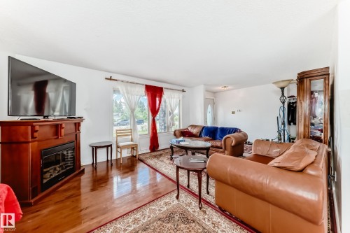 Warm and inviting living area featuring wood-finish flooring, a decorative electric fireplace with a wood-tone mantel, and a large window providing ample natural light - 6804 102A Avenue, Edmonton, AB 