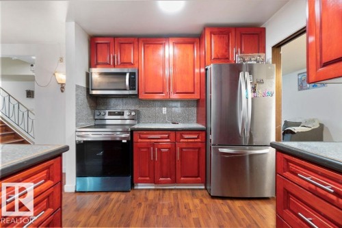 Kitchen featuring wood-finish flooring, red-stained cabinetry, stainless steel appliances including a refrigerator, range, and microwave, and a tiled backsplash - 712 Lee Ridge Road, Edmonton, AB 