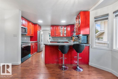 Kitchen featuring red cabinetry, wood-finish flooring, stainless steel appliances, a bar-height countertop, and multiple windows with blinds - 712 Lee Ridge Road, Edmonton, AB 