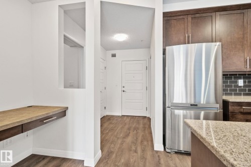 Kitchen and hallway area featuring wood-finish flooring, a built-in desk with dark wood cabinetry, a stainless steel refrigerator, dark wood kitchen cabinets, and a stone countertop - 217 2588 Anderson Way, Edmonton, AB 