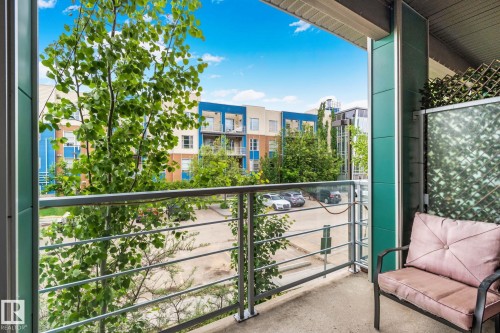 Private balcony with glass panel railing and concrete flooring, featuring vertical teal siding and an artificial foliage privacy screen - 217 2588 Anderson Way, Edmonton, AB 