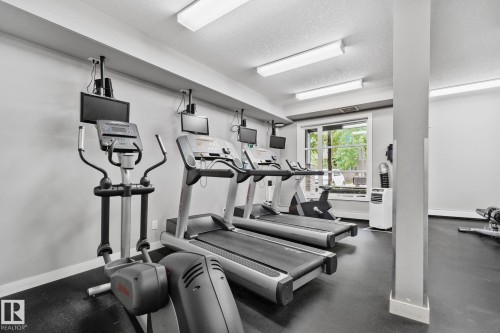 Fitness room featuring multiple cardio machines, ceiling-mounted monitors, overhead fluorescent lighting, a window with exterior views, and durable black rubber flooring - 217 2588 Anderson Way, Edmonton, AB 