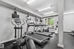 Fitness room featuring multiple cardio machines, ceiling-mounted monitors, overhead fluorescent lighting, a window with exterior views, and durable black rubber flooring - 