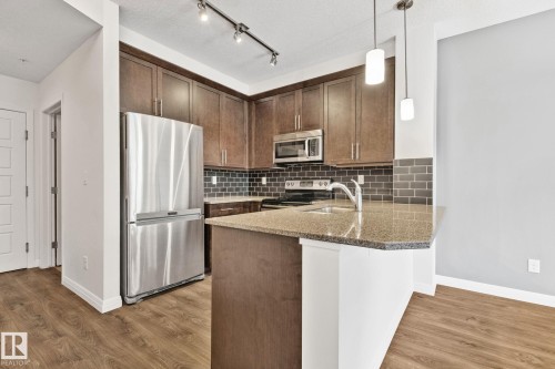 Modern kitchen featuring stainless steel appliances, dark wood-finish cabinetry, a subway tile backsplash, a peninsula with a stone-finish countertop, and wood-finish flooring - 217 2588 Anderson Way, Edmonton, AB 