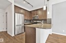 Modern kitchen featuring stainless steel appliances, dark wood-finish cabinetry, a subway tile backsplash, a peninsula with a stone-finish countertop, and wood-finish flooring - 217 2588 Anderson Way, Edmonton, AB 