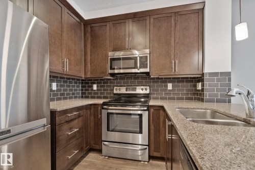 Kitchen featuring stainless steel appliances, dark wood cabinetry, a subway tile backsplash, and speckled countertops - 217 2588 Anderson Way, Edmonton, AB 