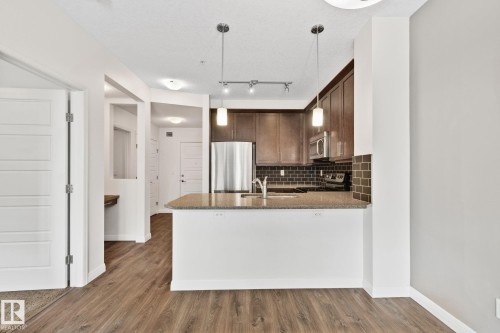 Kitchen featuring a peninsula island with an undermount sink, dark wood cabinetry, stainless steel appliances, and a dark subway tile backsplash - 217 2588 Anderson Way, Edmonton, AB 