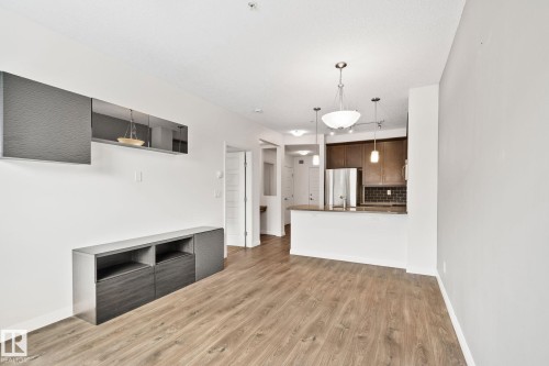 Open-concept living area with wood-finish flooring, featuring a built-in media console, an integrated kitchen with stainless steel appliances, dark wood cabinetry, and a subway tile backsplash - 217 2588 Anderson Way, Edmonton, AB 