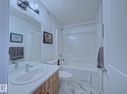 Bathroom featuring a wood-finish vanity with an integrated sink and chrome faucet, a tub/shower combination with a clear glass enclosure, and white marble-look floor tiles - 