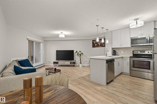 Open-concept living space featuring wood-finish flooring, a kitchen island with pendant lighting, stainless steel appliances, white cabinetry, and a sliding glass door to an outdoor area - 210 2229 44 Avenue, Edmonton, AB 