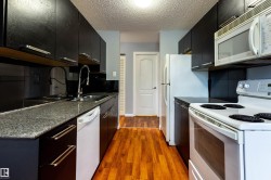 Galley kitchen featuring dark cabinetry, granite-finish countertops, wood-finish flooring, a double basin sink with a gooseneck faucet, and a full suite of white appliances including a refrigerator, range, microwave, and dishwasher - 