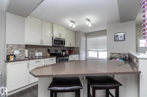 Kitchen featuring a peninsula island with counter seating, light-toned cabinetry, stainless steel appliances, a mosaic tile backsplash, and a window with blinds - 46 4050 Savaryn Drive, Edmonton, AB 