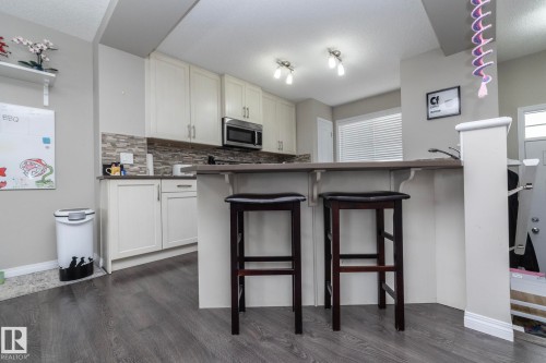 Kitchen featuring wood-finish flooring, white cabinetry, stainless steel microwave, stone backsplash, and a breakfast bar - 46 4050 Savaryn Drive, Edmonton, AB 