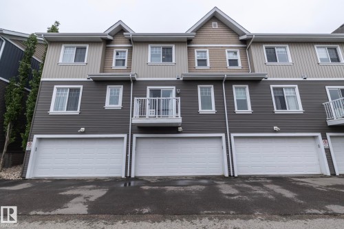 Multi-level exterior featuring grey horizontal siding, tan shake accents, white-trimmed windows, and three individual garages with white panel doors - 46 4050 Savaryn Drive, Edmonton, AB 